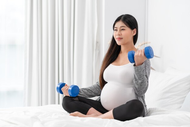 La mujer embarazada feliz asiática está sentada y ejercita en la cama. concepto de embarazo, maternidad, personas y expectativa