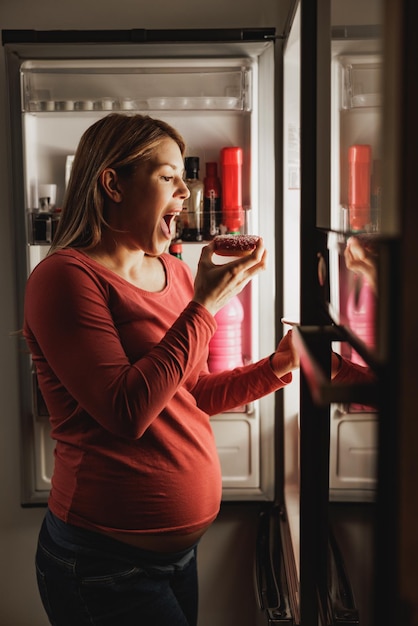 Foto mujer embarazada feliz comiendo donut frente al refrigerador abierto en la cocina por la noche.