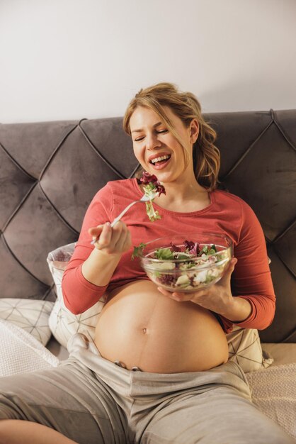 Mujer embarazada feliz comiendo ensalada de verduras frescas mientras se sienta en la cama en su dormitorio.