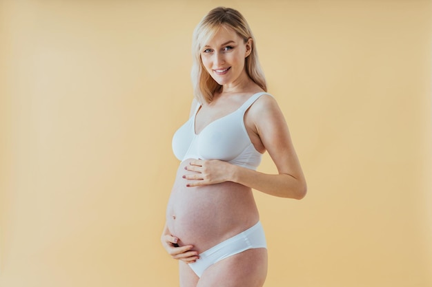 Mujer embarazada feliz con gran barriga sobre fondo de color - Mujer joven con ropa interior esperando un bebé - Embarazo, maternidad, personas y concepto de expectativa -