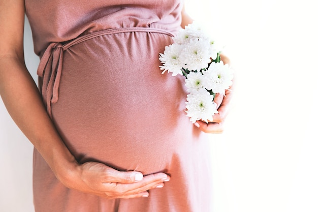 Mujer embarazada con flores de crisantemos tiene las manos sobre el vientre en fondo blanco.