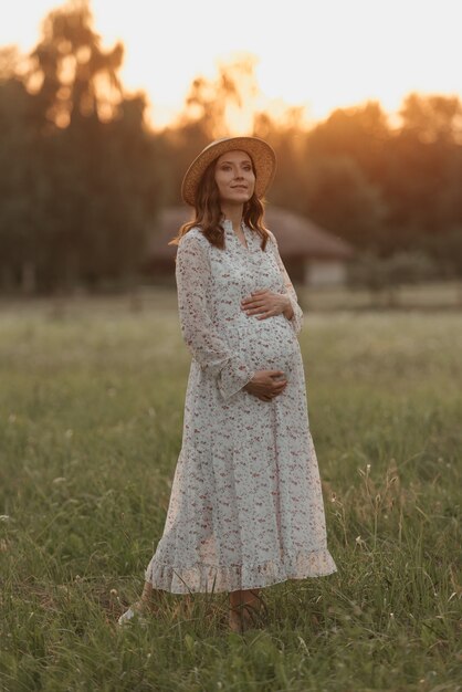 Una mujer embarazada en un fondo de puesta de sol está posando y sosteniendo su vientre con las manos cerca de la granja