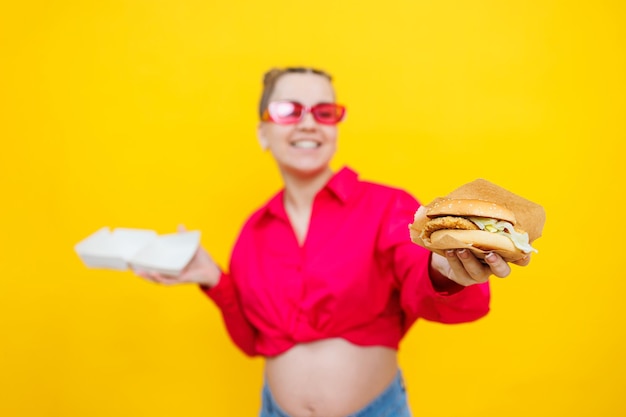 Mujer embarazada hambrienta sosteniendo una hamburguesa comiendo comida chatarra posando sobre fondo amarillo en el estudio Mujer disfrutando de una gran hamburguesa El concepto de comer poco saludable y comer en exceso durante el embarazo