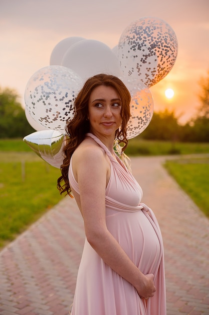 Mujer embarazada hermosa feliz en el vestido largo rosado que camina con los globos de aire en el p verde