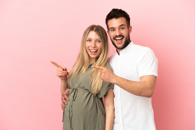 Mujer embarazada y hombre sobre fondo rosa aislado apuntando con el dedo hacia un lado y presentando un producto