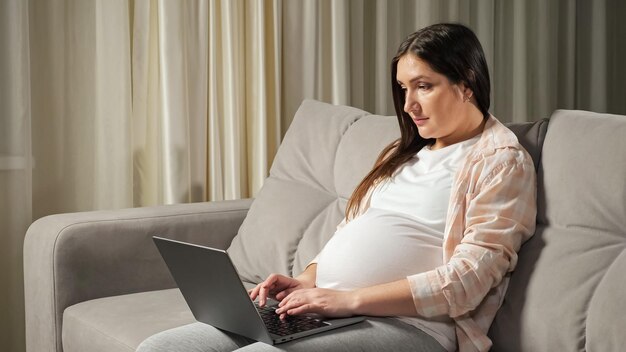 Mujer embarazada independiente trabaja a través de una computadora portátil desde casa en el sofá