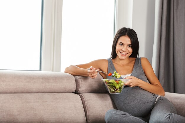 Mujer embarazada en el interior de su casa sentado en el sofá comer ensalada.