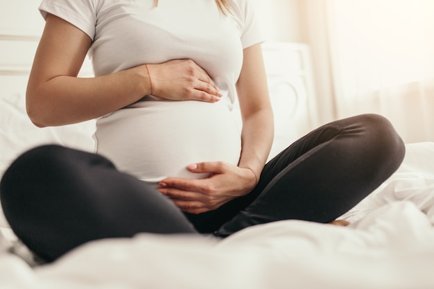 Mujer embarazada irreconocible tocando el vientre y sentado con las piernas cruzadas en una cómoda cama en casa