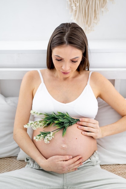 Una mujer embarazada en jeans con el vientre abierto y una flor lo acaricia con las manos La futura madre está esperando y preparándose para el nacimiento de un niño en casa en un dormitorio luminoso