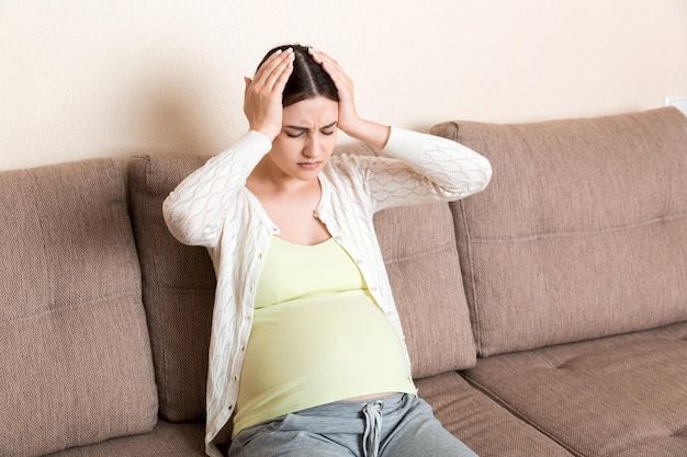 Mujer embarazada joven con dolor de cabeza sentada en el sofá Concepto de expectativa de embarazo
