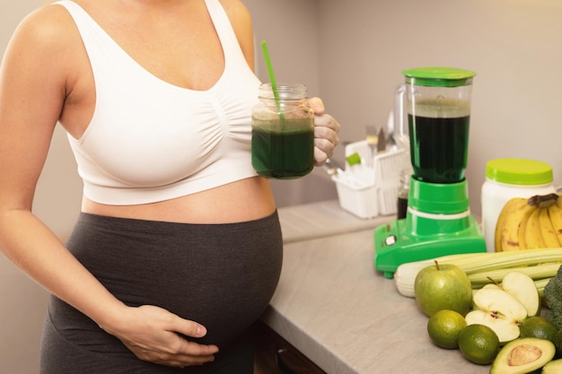 Mujer embarazada joven y feliz bebiendo batido verde saludable en casa