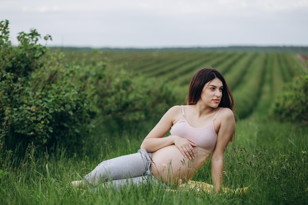 Mujer embarazada joven feliz que se sienta en la hierba que hace yoga en día de verano