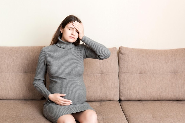 Mujer embarazada joven que sufre de dolor de cabeza en el sofá en el concepto de atención médica de maternidad en el hogar