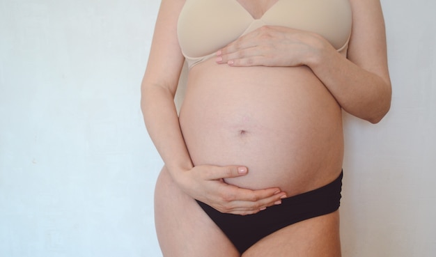 Una mujer embarazada en lencería se encuentra sobre un fondo de una pared blanca y acaricia su vientre. Concepto: maternidad y embarazo.