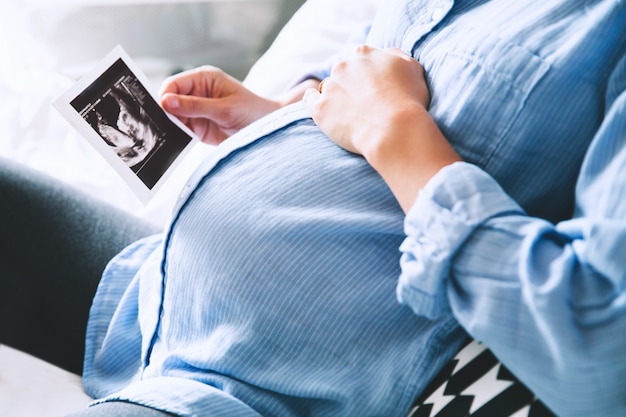 Mujer embarazada manteniendo la mano sobre el vientre y sosteniendo una imagen de ultrasonido en el interior de las casas. Concepto de embarazo, paternidad, preparación y expectativa. Primer plano, copie el espacio, en el interior.