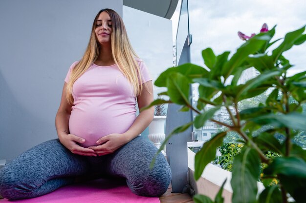 Mujer embarazada meditando sobre una colchoneta tocándose el vientre con una planta en primer plano