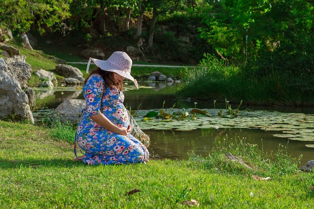 Mujer embarazada mirando su vientre arrodillado sobre el césped cerca del lago
