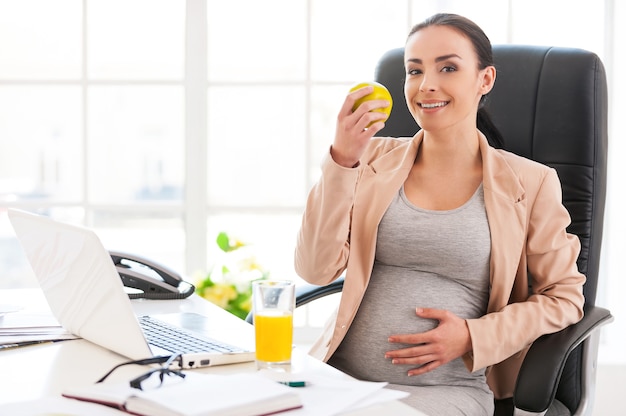 Mujer embarazada en la oficina. Hermosa empresaria embarazada comiendo manzana y sonriendo a la cámara mientras está sentada en su lugar de trabajo