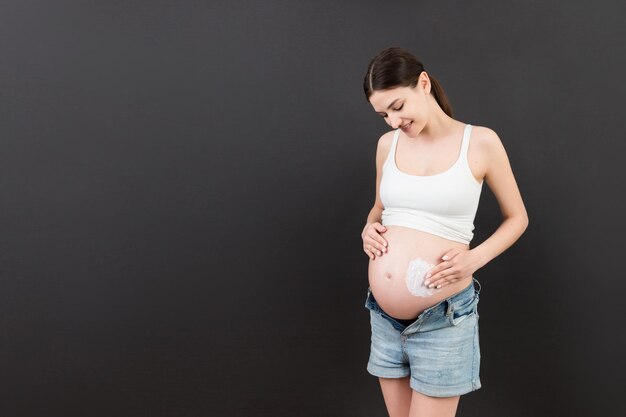 mujer embarazada con pantalones vaqueros abiertos aplicando loción hidratante en su vientre para evitar estrías en un fondo colorido con espacio de copia Cuidado de la piel durante el embarazo concepto