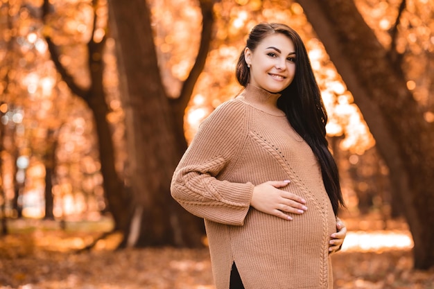Mujer embarazada de pie en el bosque del parque de la ciudad de otoño, sosteniendo acariciando su vientre redondo con el bebé