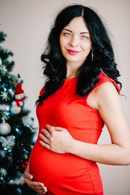 mujer embarazada posando junto al árbol de Navidad en casa. ¡Feliz Navidad y Felices Fiestas! Concepto de embarazo, vacaciones, personas y expectativa.