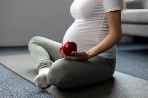 Mujer embarazada practicando yoga, sosteniendo manzana roja en casa. Embarazo, concepto de estilo de vida saludable