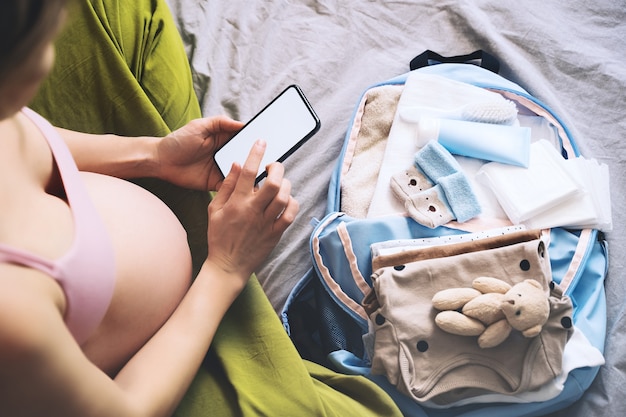 Mujer embarazada preparándose para el trabajo de embalaje para el hospital haciendo notas en el teléfono inteligente