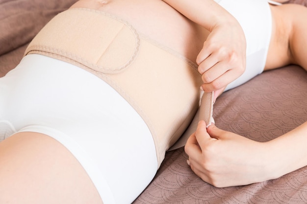 Mujer embarazada de primer plano usa vendaje en el vientre en casa en la cama cinturón de apoyo abdominal ortopédico