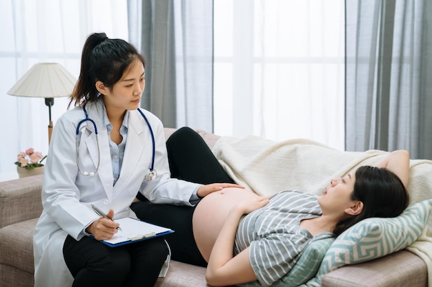 Mujer embarazada a la que el médico le da un control prenatal en la sala de estar del hogar en el sofá. matrona femenina escribiendo en el portapapeles con una mano tocando el elegante vientre de la dama de maternidad en el sofá en un examen regular