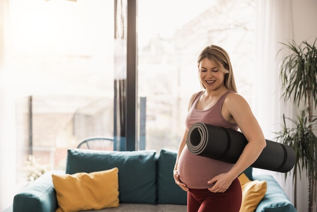 La mujer embarazada que sostiene la colchoneta de ejercicios está lista para practicar yoga en casa por la mañana.