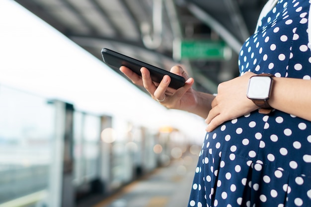 Foto mujer embarazada que usa el teléfono móvil mientras espera el autobús o el tren de cercanías.