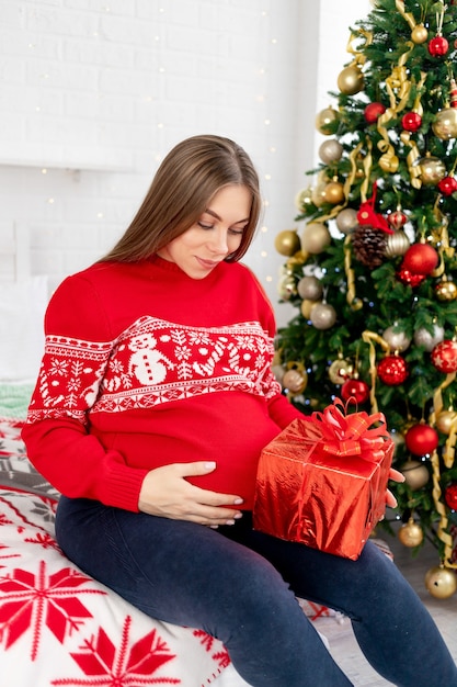 Una mujer embarazada con un regalo en un suéter rojo debajo del árbol de Navidad en casa está acariciando su gran barriga y soñando con un bebé y disfrutando del año nuevo y la Navidad.