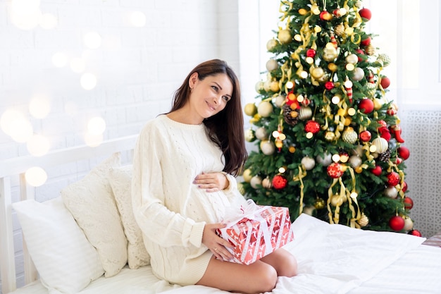 La mujer embarazada se regocija y mira los regalos de Navidad en el dormitorio de su casa sobre el fondo del árbol de Navidad