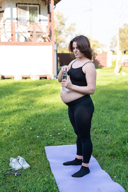 Mujer embarazada en ropa deportiva bebiendo una botella de agua después de hacer  yoga al aire libre tomando un descanso en la práctica de yoga  acondicionamiento físico y meditación concepto de salud