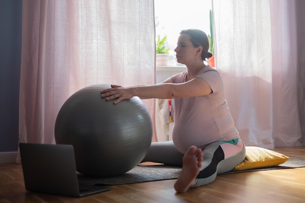 Mujer embarazada en ropa deportiva, haciendo yoga, entrenamiento de pilates, postura de ángulo amplio sentado hacia adelante, Upavishtha Konasana en casa