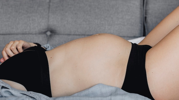 Mujer embarazada en ropa interior negra acostada en un sofá gris
