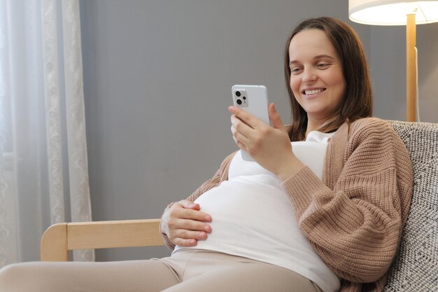 Mujer embarazada satisfecha con un teléfono inteligente sentada en el sofá en casa usando Internet en su teléfono móvil madre embarazada haciendo una llamada de video haciendo selfie mensajería en la aplicación móvil de mensajes de texto