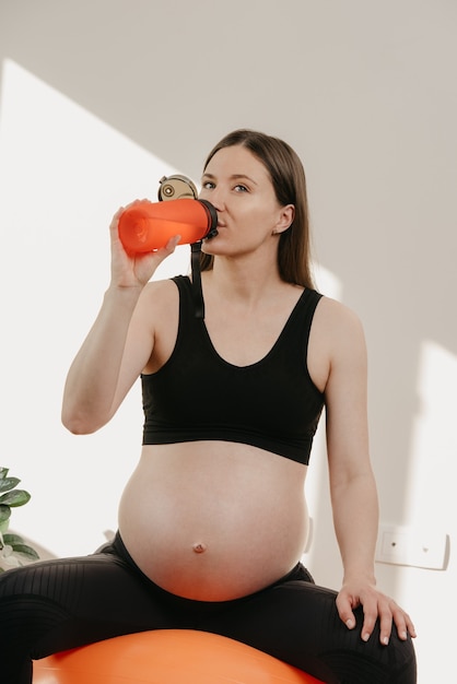 Una mujer embarazada sentada sobre una pelota de ejercicios naranja y bebiendo agua de