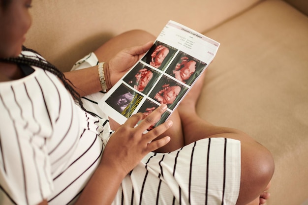Mujer embarazada sentada en el sofá de casa y mirando la imagen de ultrasonido de su bebé