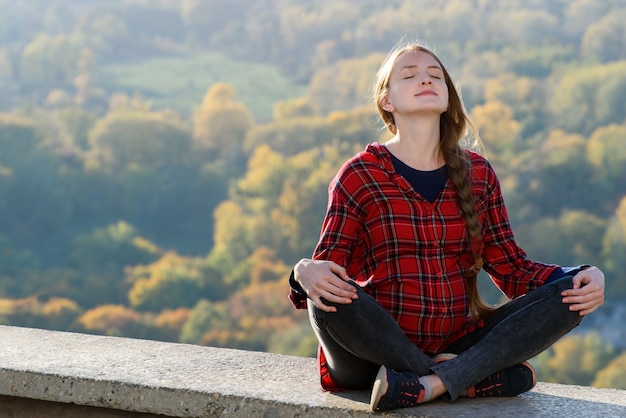 La mujer embarazada se sienta en una colina con los ojos cerrados. Meditación. Bosque de otoño en el espacio