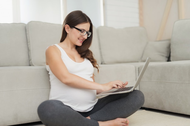 La mujer embarazada se sienta con las piernas cruzadas y trabaja en la computadora portátil gris.