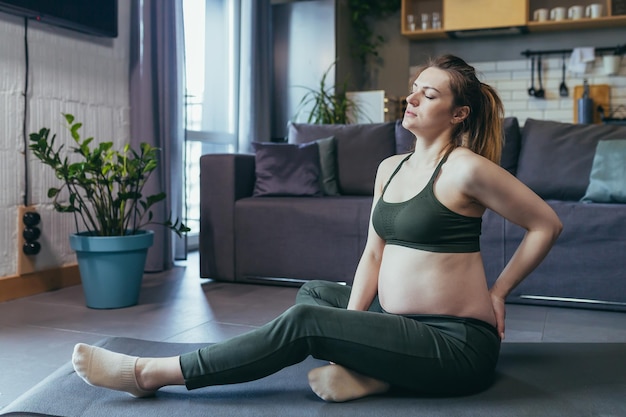 Una mujer embarazada siente dolor en el cuerpo después de hacer ejercicio sosteniendo su pierna hacia atrás Sentada en casa en una alfombra deportiva