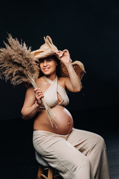 Mujer embarazada con un sombrero de paja con ropa beige con un ramo en sus manos en el estudio sobre un fondo negro.