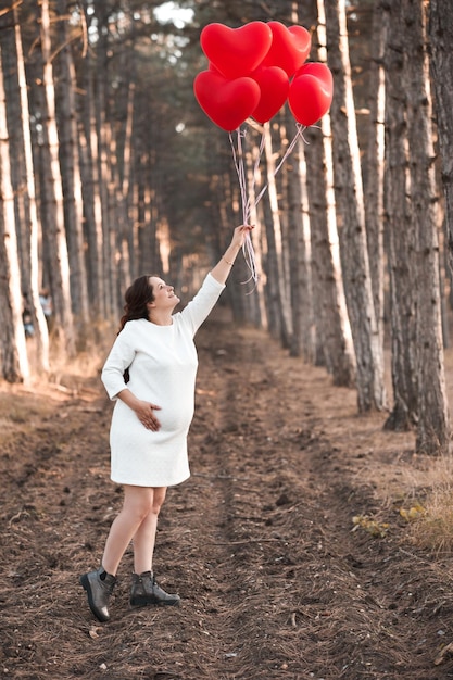 Foto mujer embarazada sonriente de 25 a 30 años sosteniendo globos de corazón rojo con vestido blanco posando al aire libre