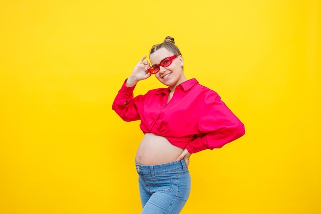 Una mujer embarazada sonriente con una camisa de algodón rosa y jeans sostiene suavemente su vientre embarazado Posando para la cámara Concepto de embarazo fácil y feliz