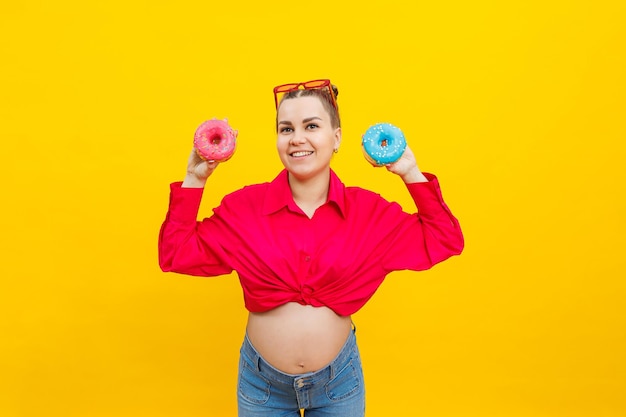 Una mujer embarazada sonriente con una camisa rosa sostiene rosquillas en sus manos sobre un fondo amarillo Alimentos dulces durante el embarazo Alimentos nocivos durante el embarazo