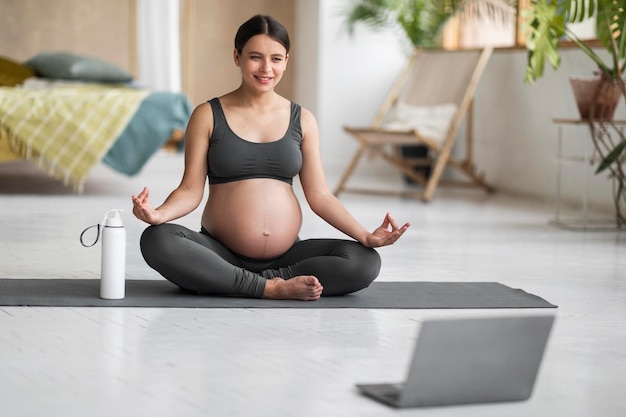 Mujer embarazada sonriente con laptop practicando yoga en línea en casa