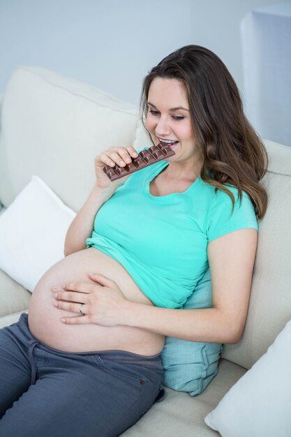 Mujer embarazada sonriente que come el chocolate en el sofá