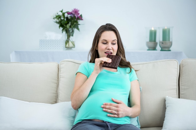 Mujer embarazada sonriente que come el chocolate en el sofá