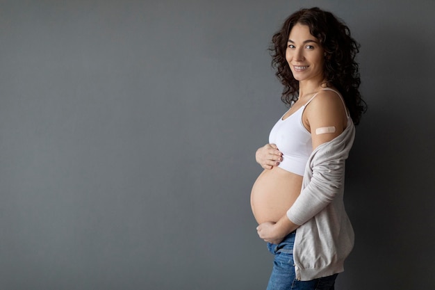 Mujer embarazada sonriente que muestra el brazo con vendaje adhesivo después de la inyección de la vacuna